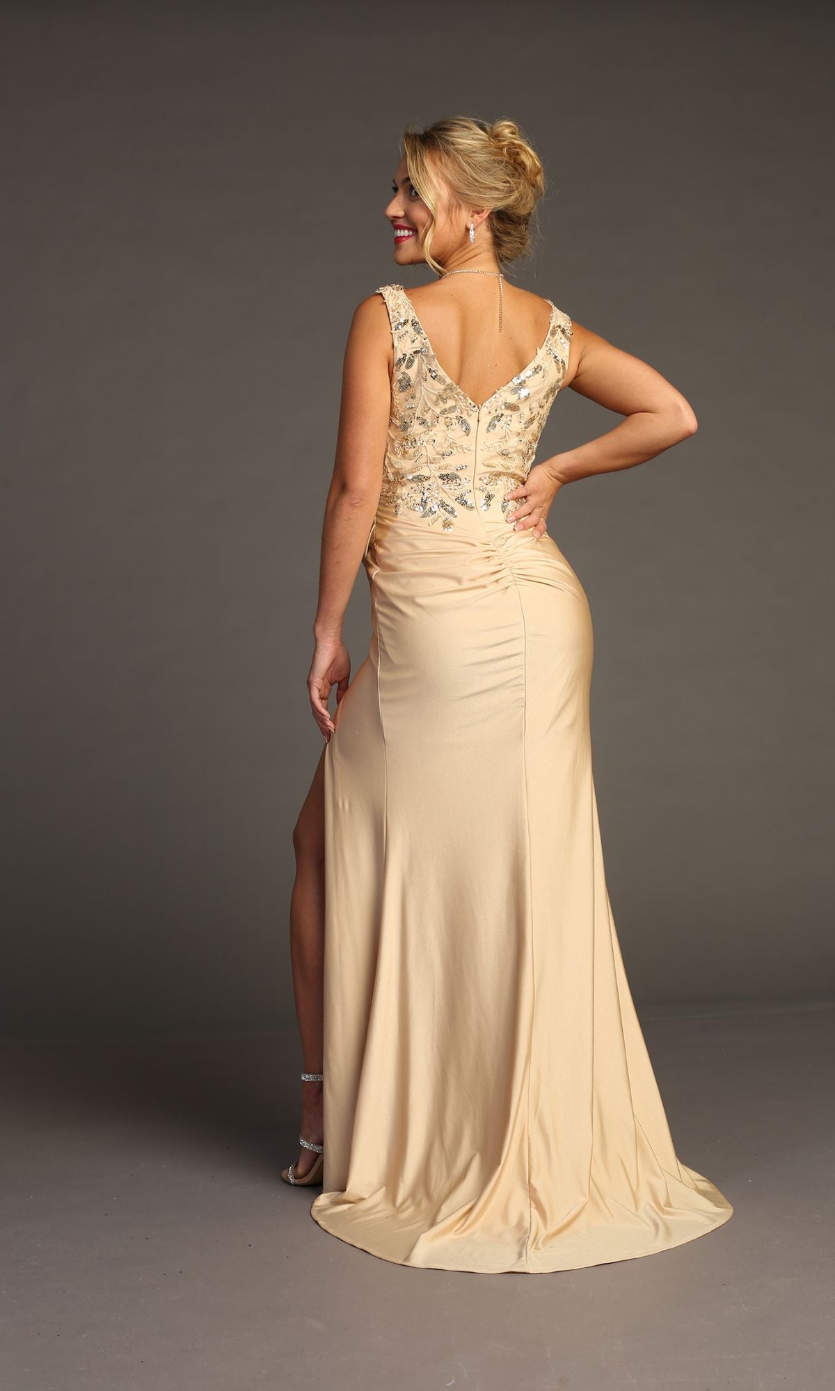 A woman stands facing away in the Sequin-Bodice Long Prom Dress: Chicas YG5032, featuring silver sequin embellishments on top and a side slit, ideal as a formal or prom dress, against a plain studio backdrop.